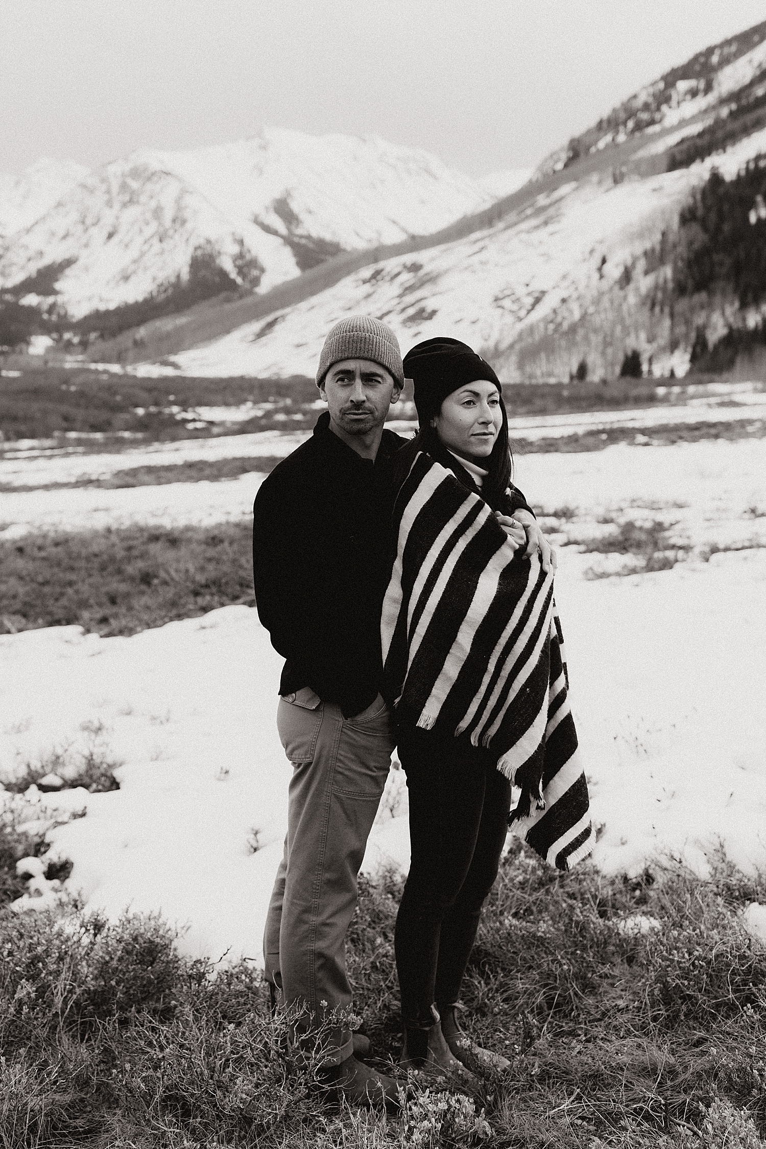 man and woman wrapped in blanket by mountain showing 3 tips for engagement photos in the snow