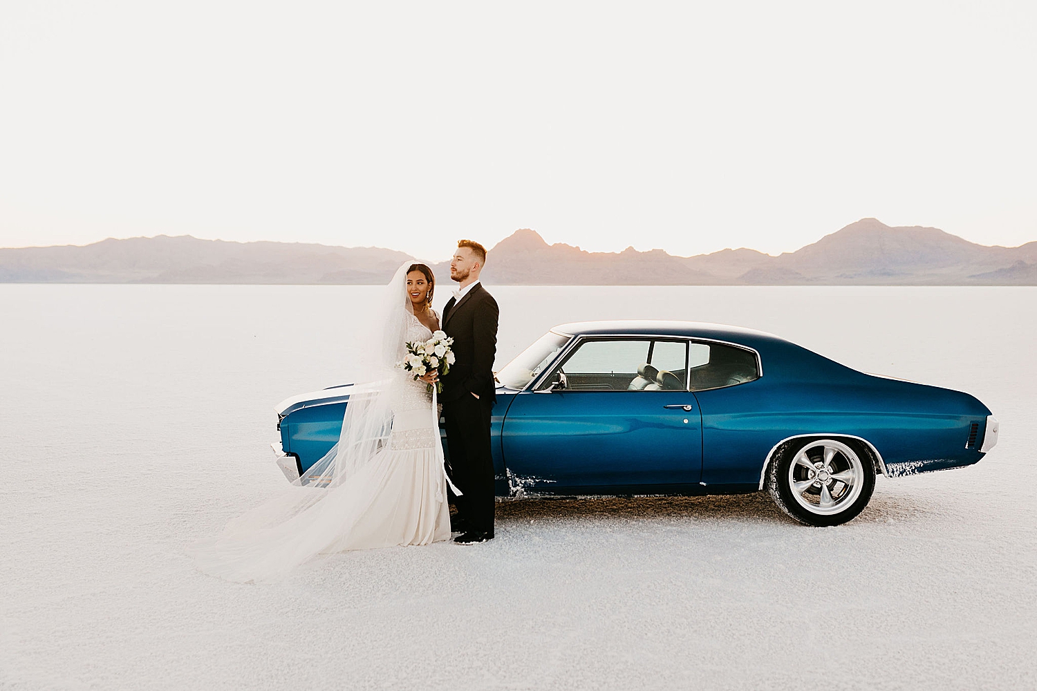 woman with veil and man in suit stand next to blue sports car by Destination wedding photographer