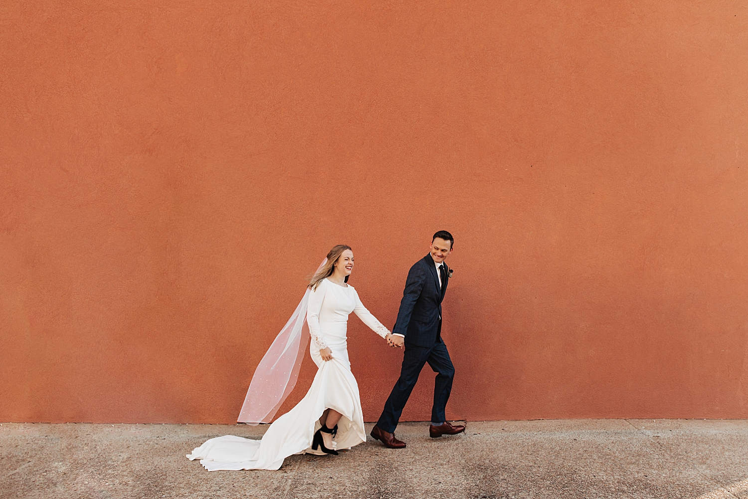 bride and groom walk up the street in front of orange wall for sunrise shoot