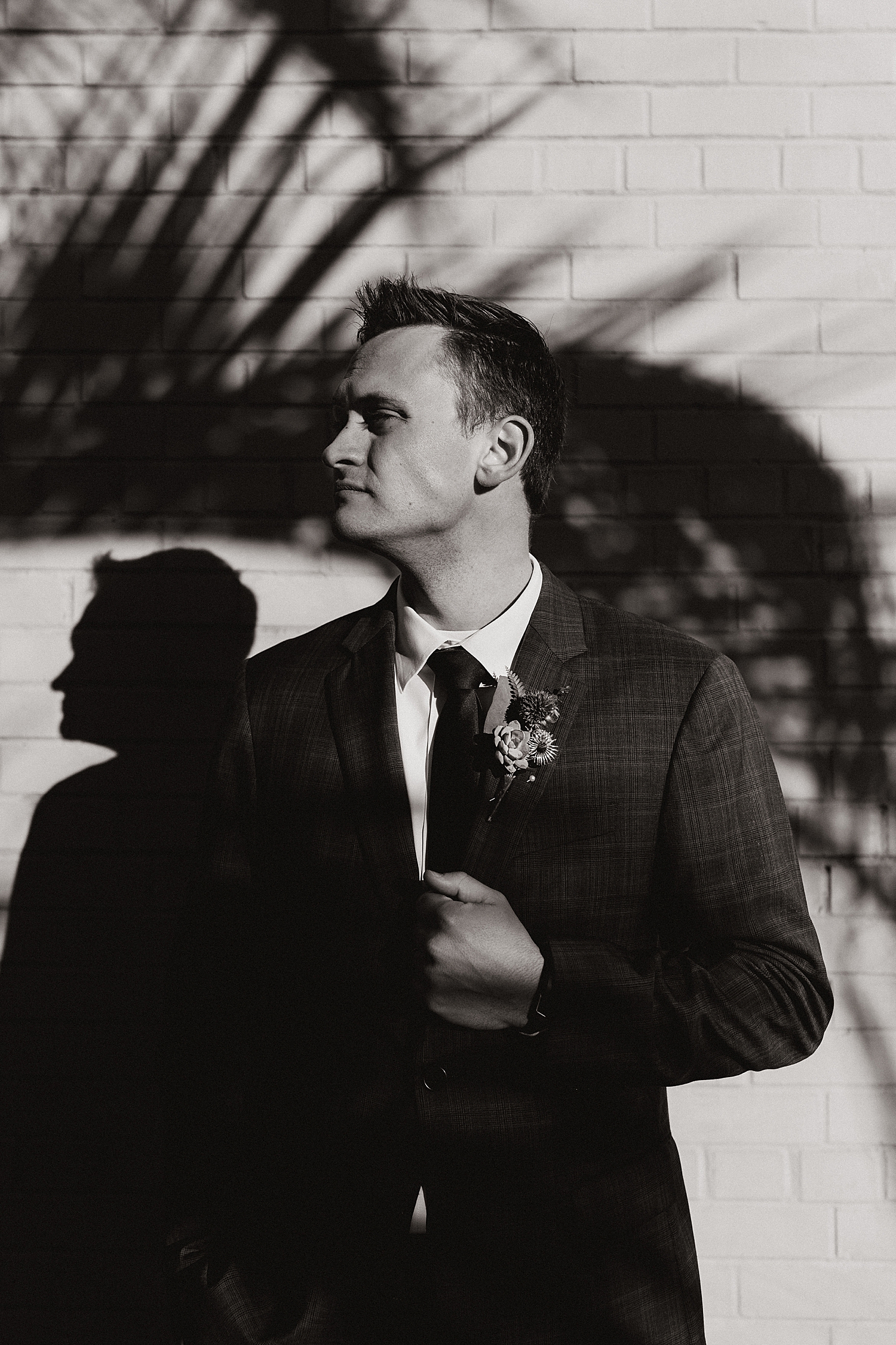 groom stands in front of brick wall under palm tree shadows for sunrise shoot by Destination wedding photographer