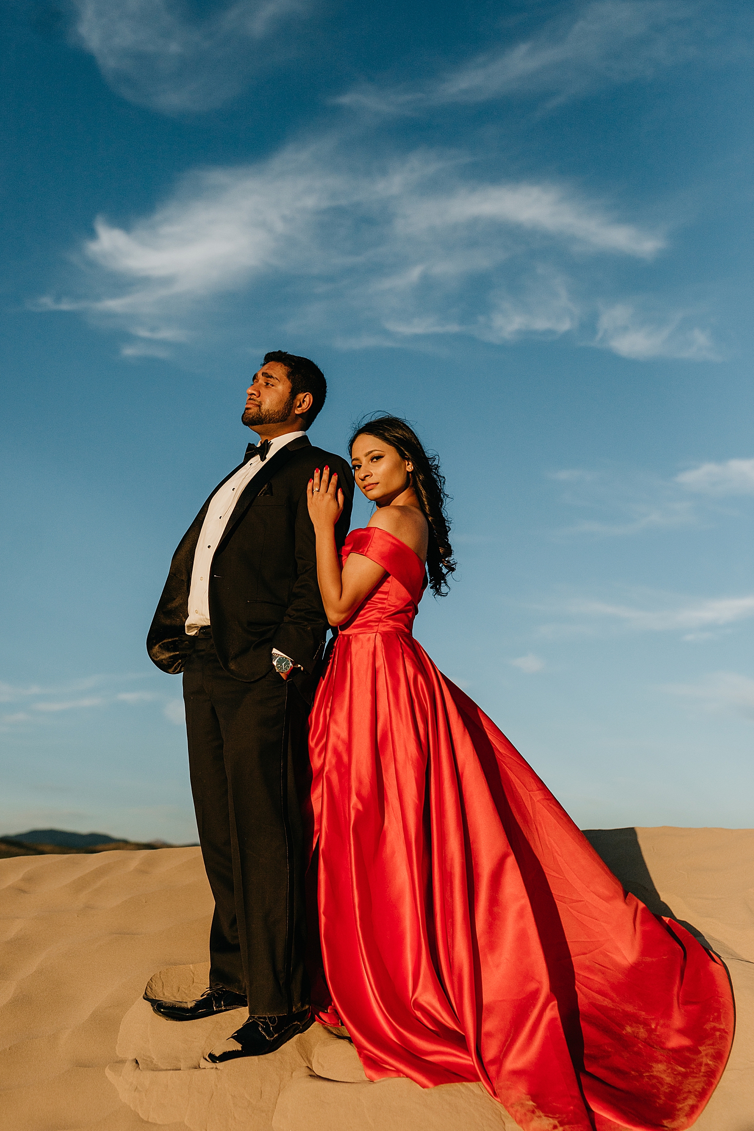 engaged couple in formal wear stand on hill under blue skies for Sand Dune engagement session