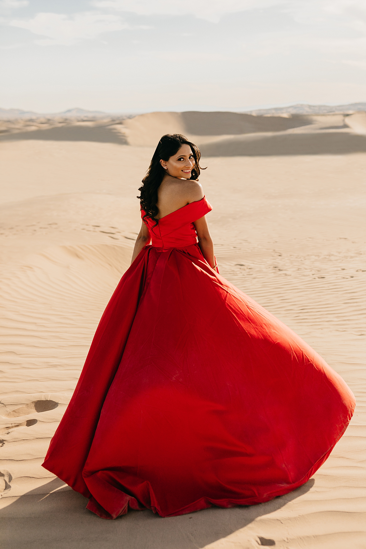brunette in bright red ballgown looks over her shoulder for Sand Dune engagement session