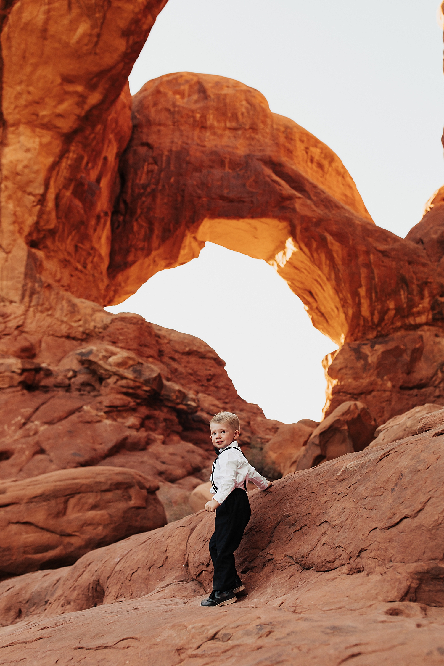 little toddler in suit stands near rock under an arch by Destination elopement photographer