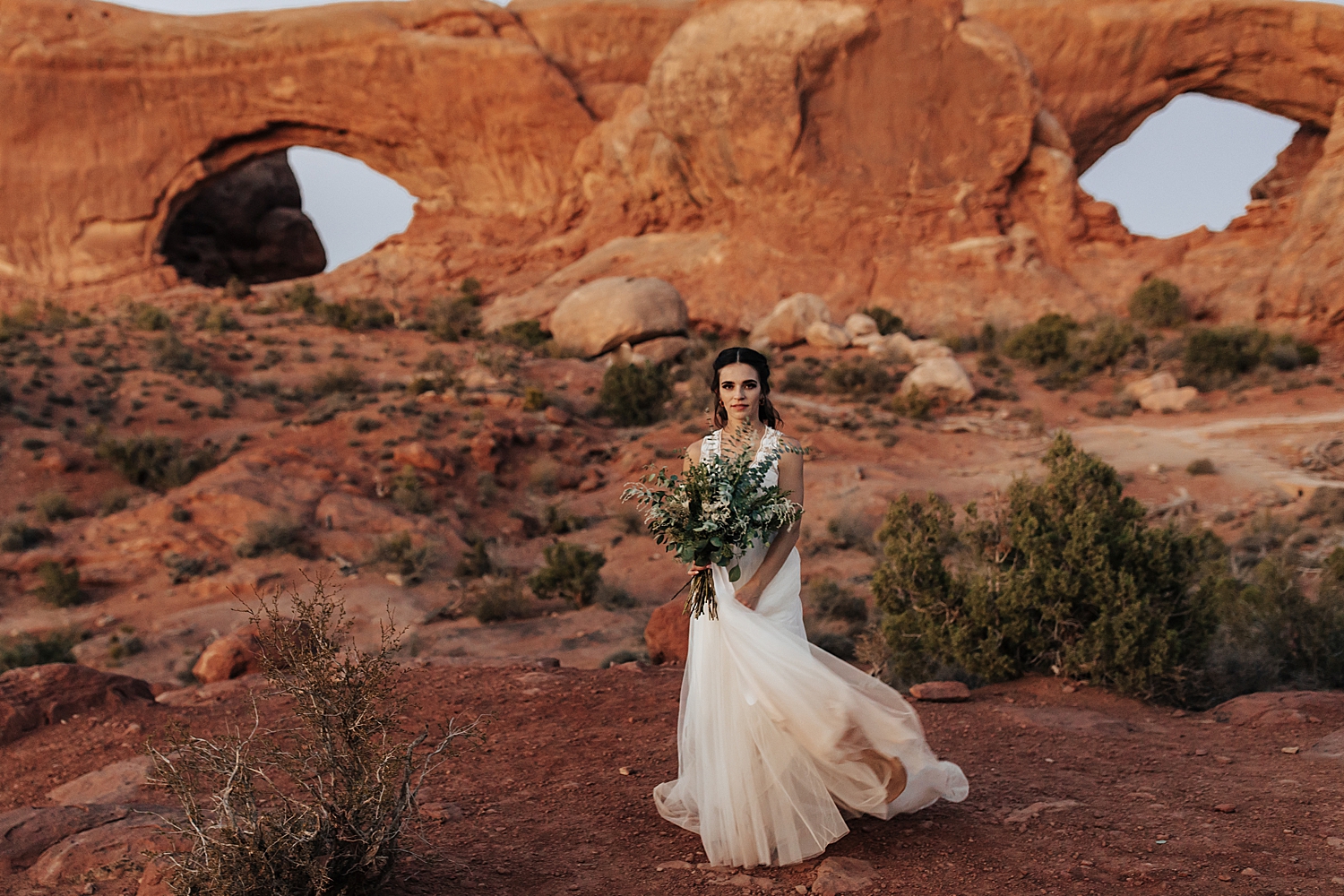 brunette in long white dresses walks in front of arches by Destination elopement photographer