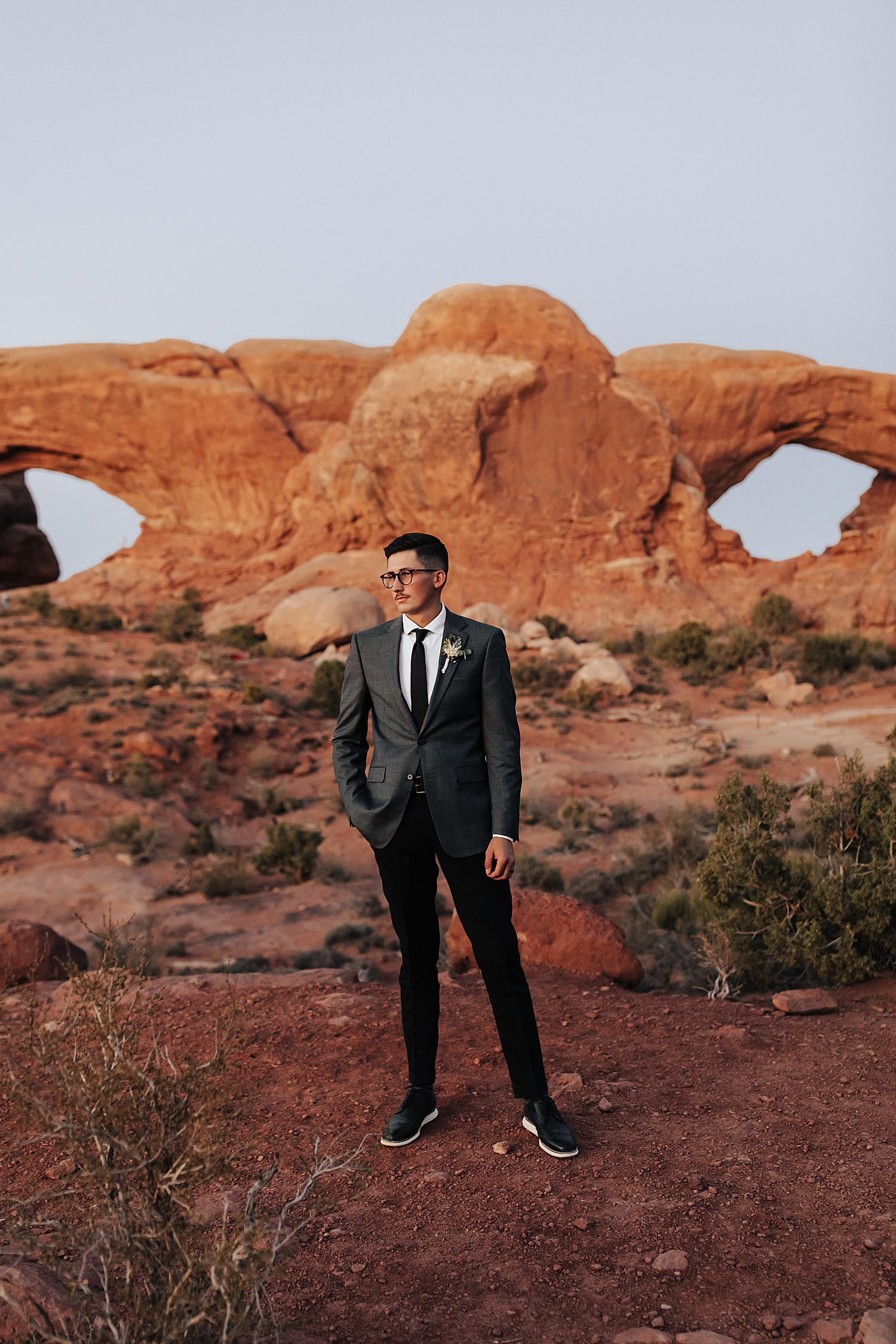 groom in black suit looks off at the sunset in front of red rocks by Nicole Aston Photography