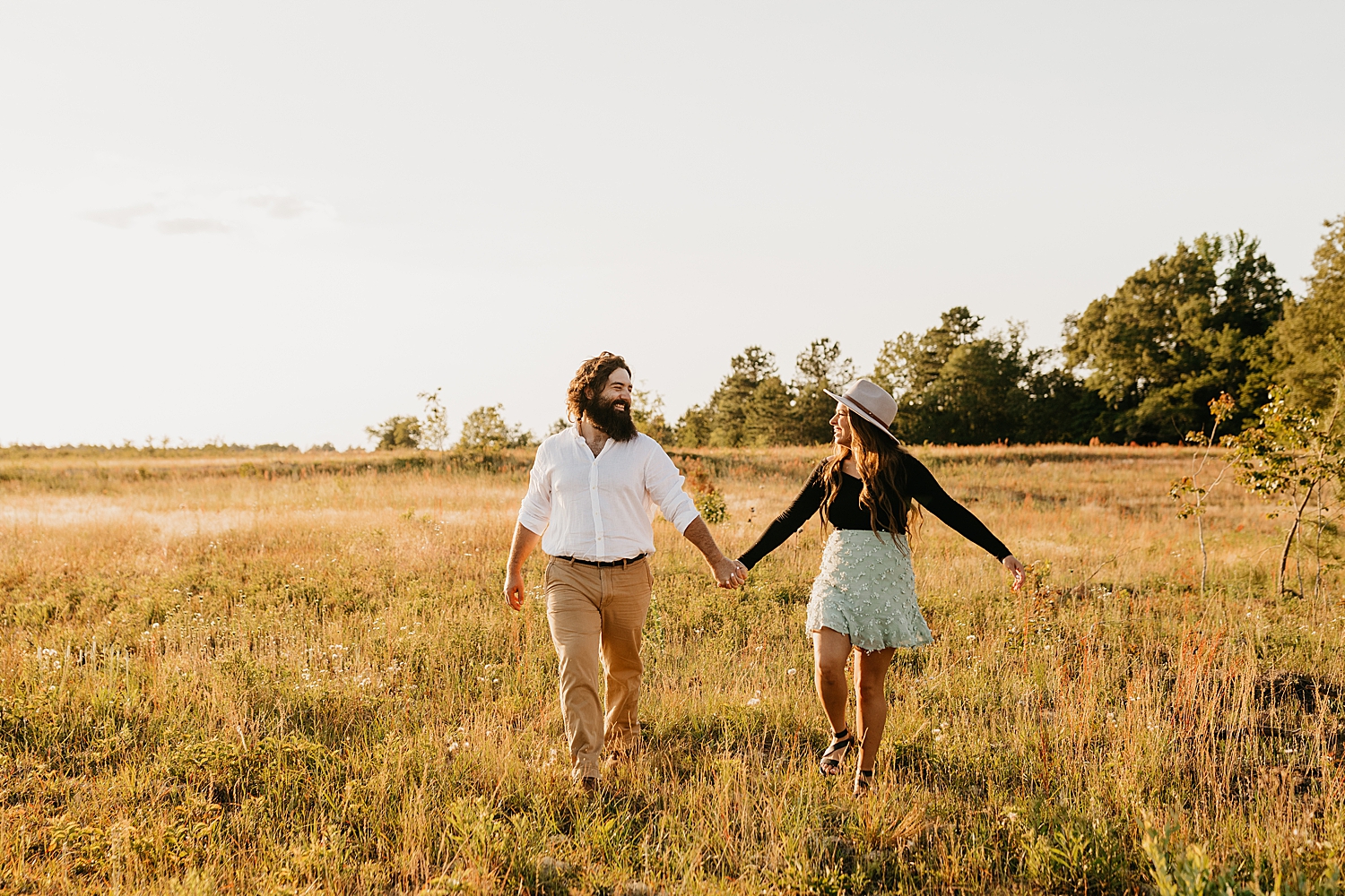 man in white shirt walks with woman in skirt in field by destination wedding photographer