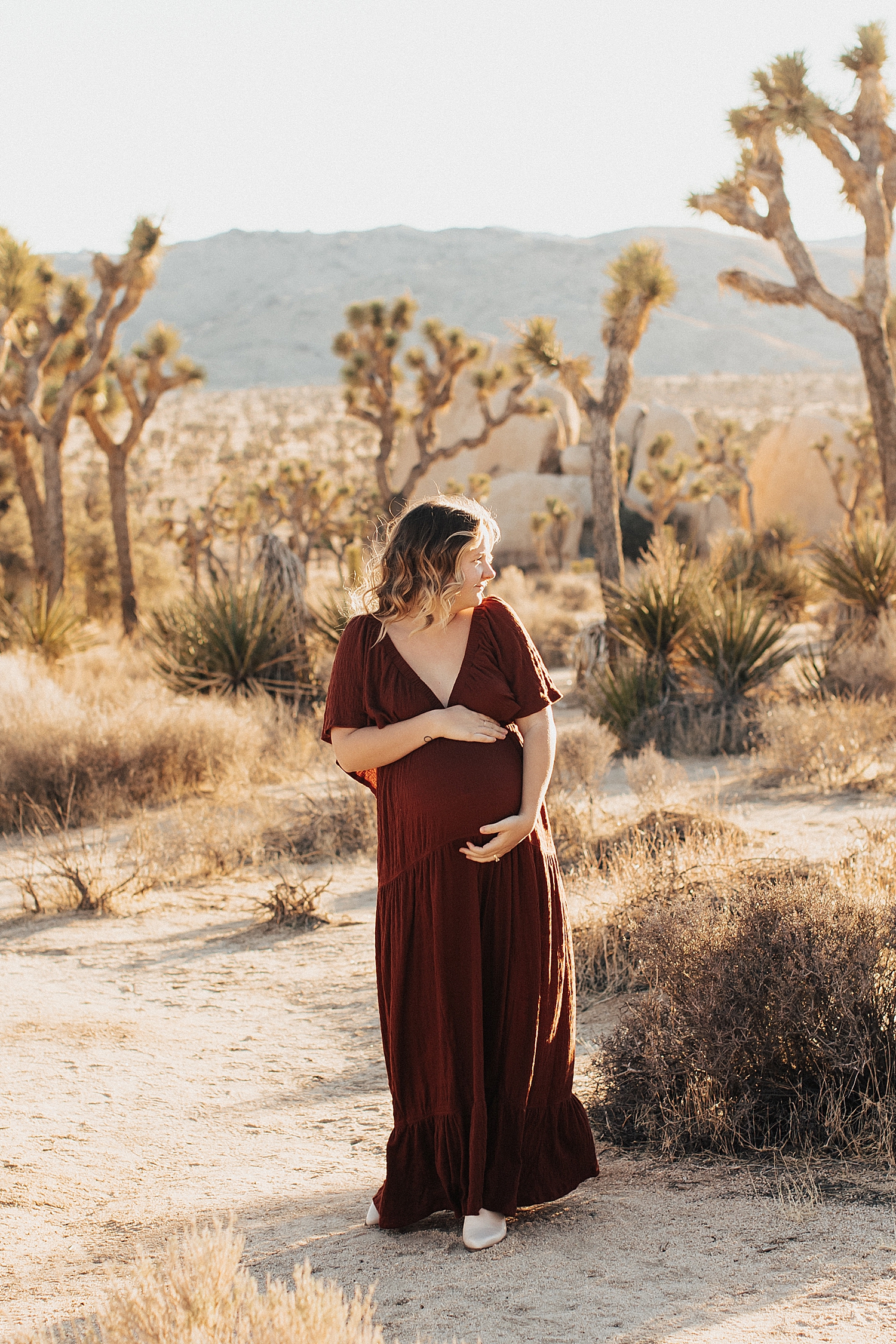 mom in maroon dress in desert holding pregnant belly by Nicole Aston Photography