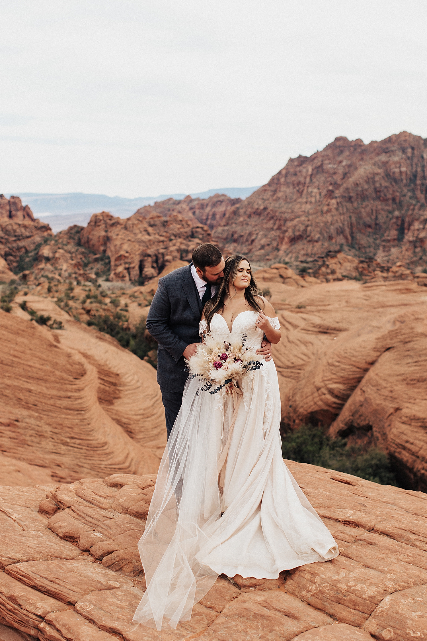 man leans in to kiss his new wife's neck standing on red rocks at Snow Canyon
