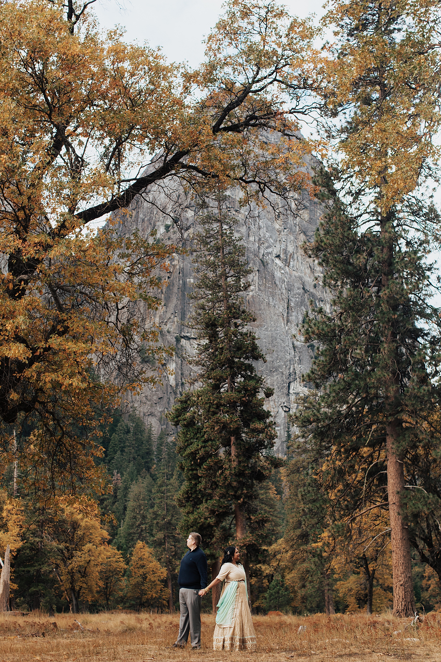 couple hold hands, standing back to back, with large boulder in the distance for Yosemite engagement session