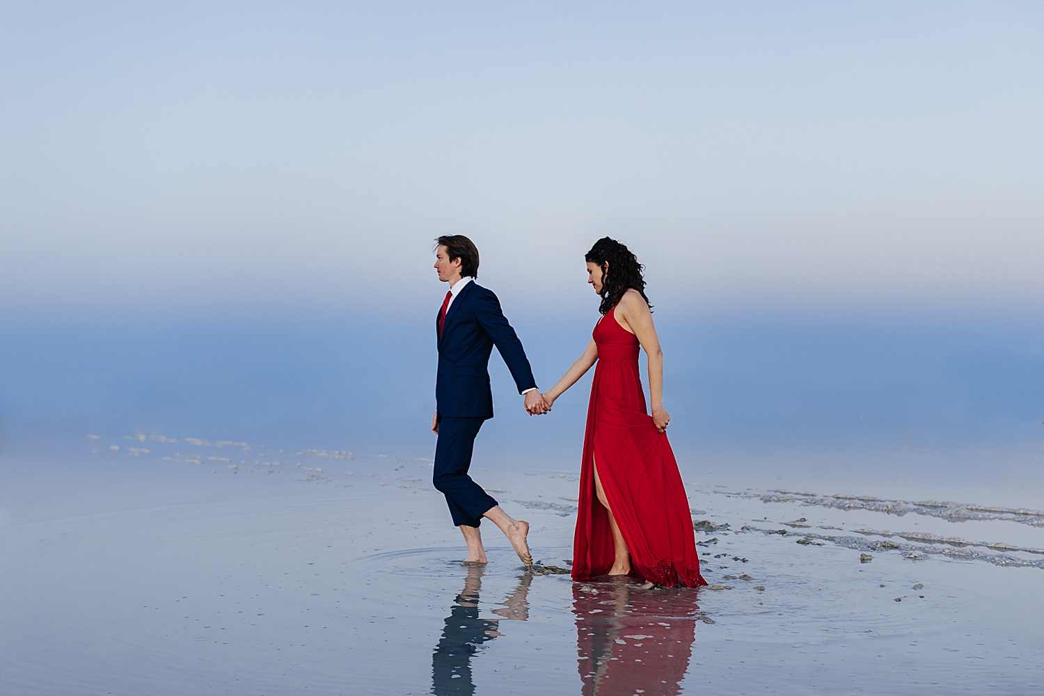 brunette in long red dress walks hand in hand with her groom over the flooded salt flats