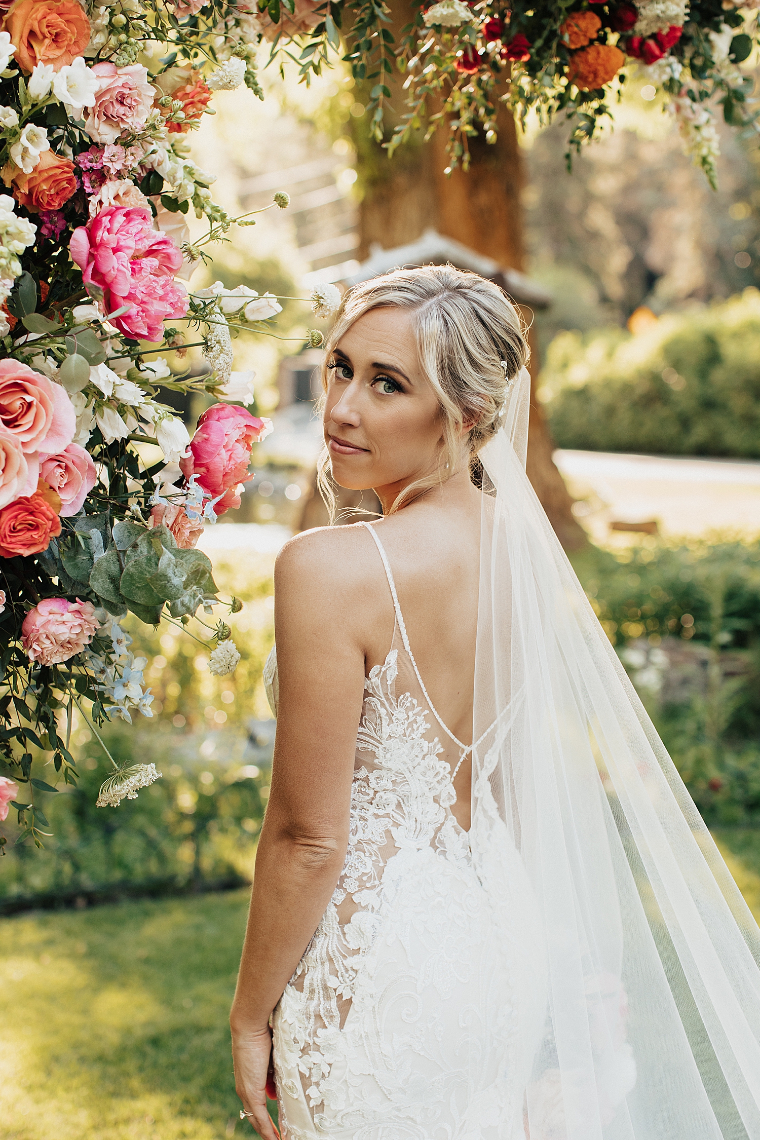 blonde bride stands under floral arch  looking over shoulder by Nicole Aston Photography