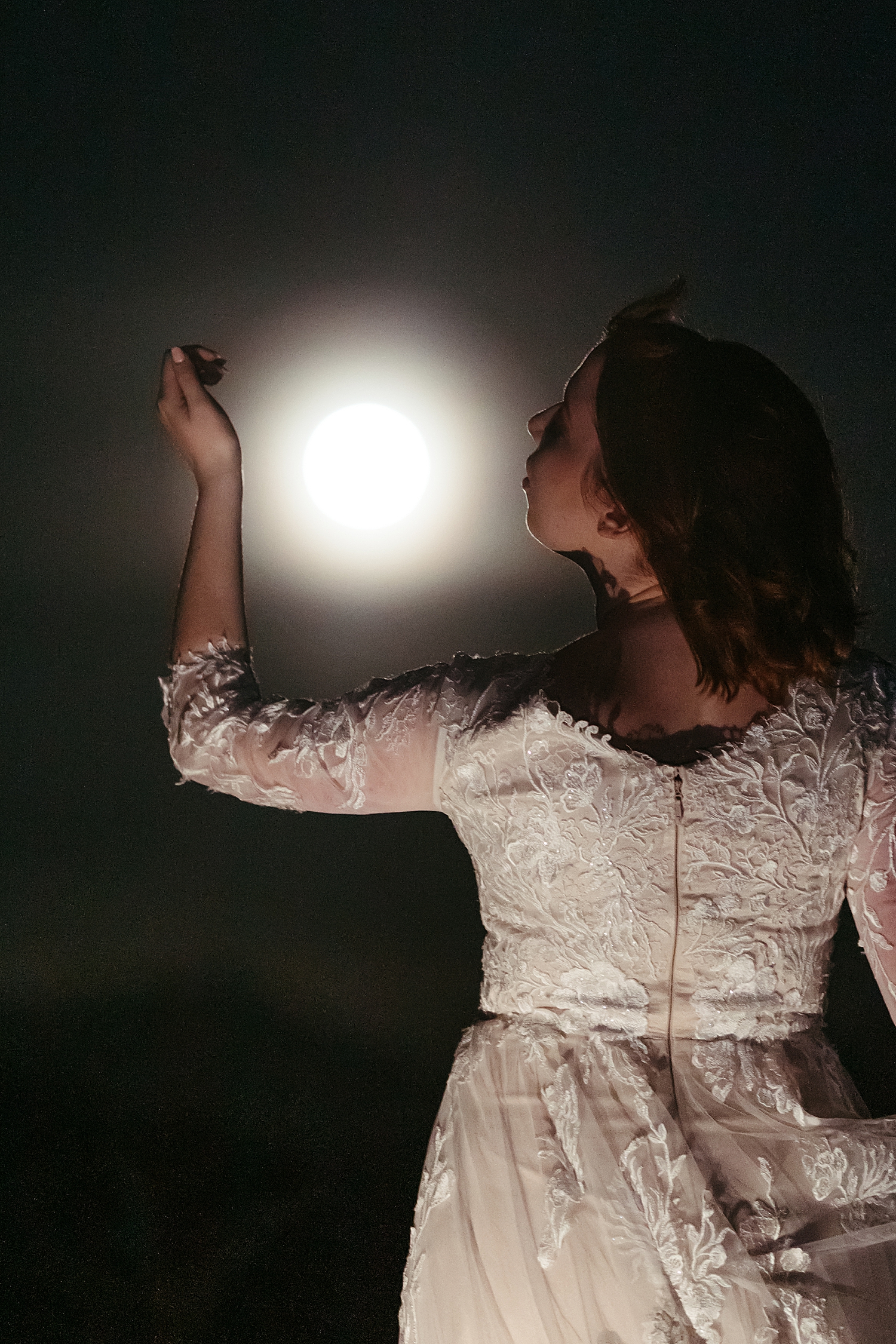 bride holds her arm up around the moon in the dark sky for full moon bridal session