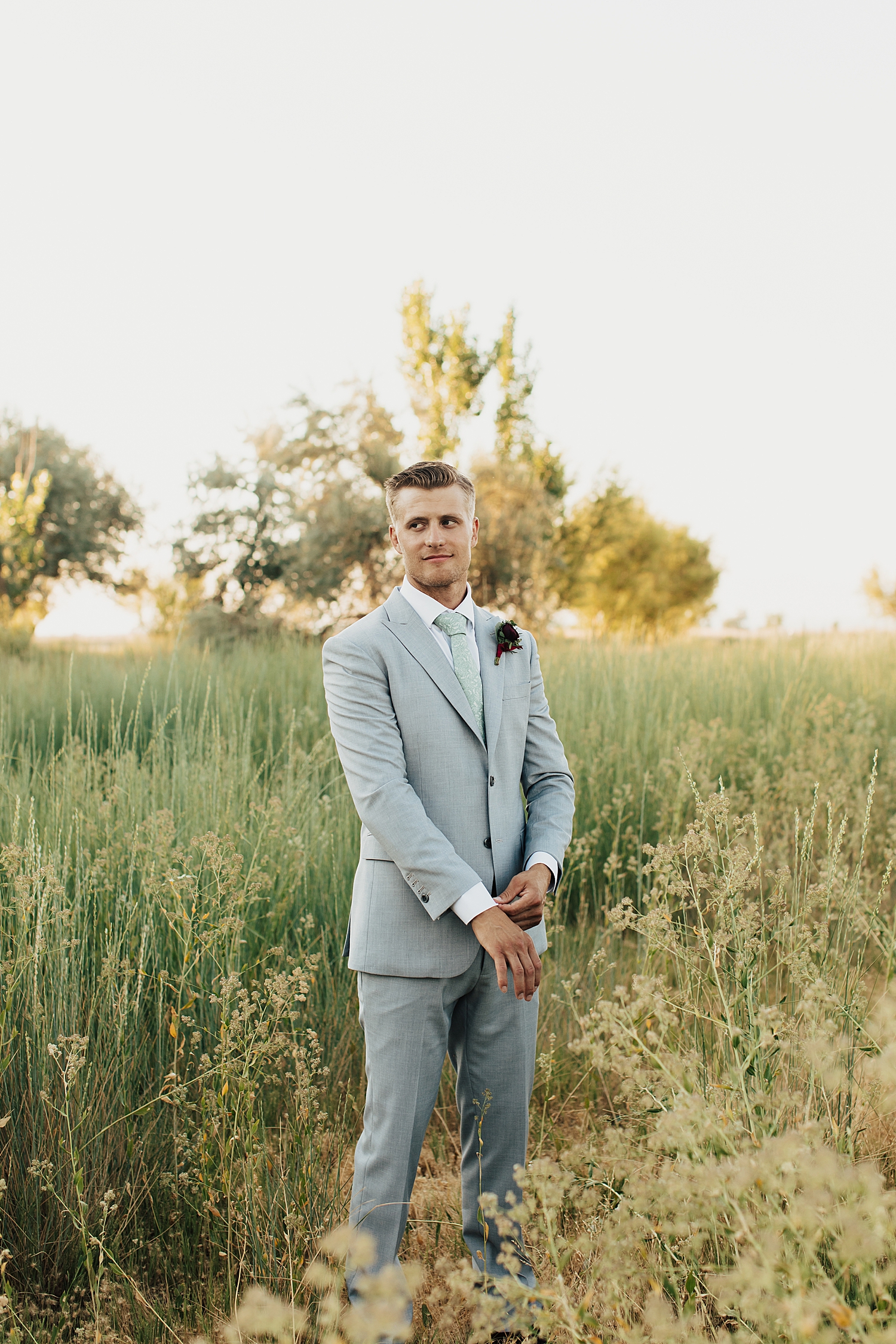 groom in gray suit adjusts sleeves in field at bountiful pond