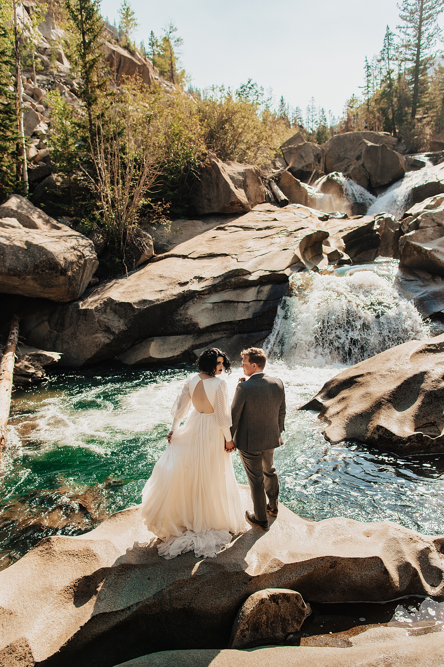 bride and groom stand on rock overlooking blue waterfall by destination wedding photographer
