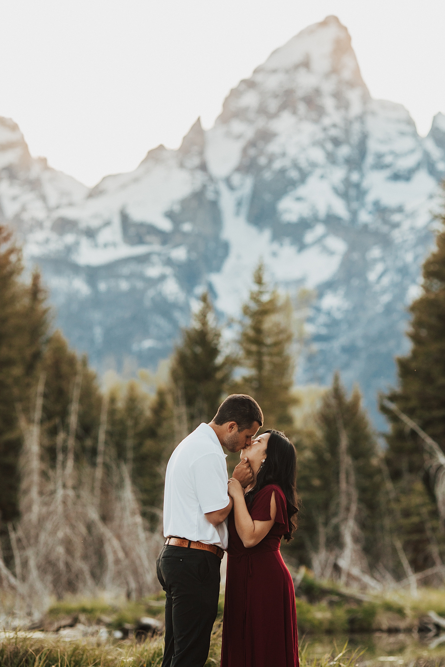 woman in red dress kisses man in white shirt in front of mountain by Nicole Aston Photography