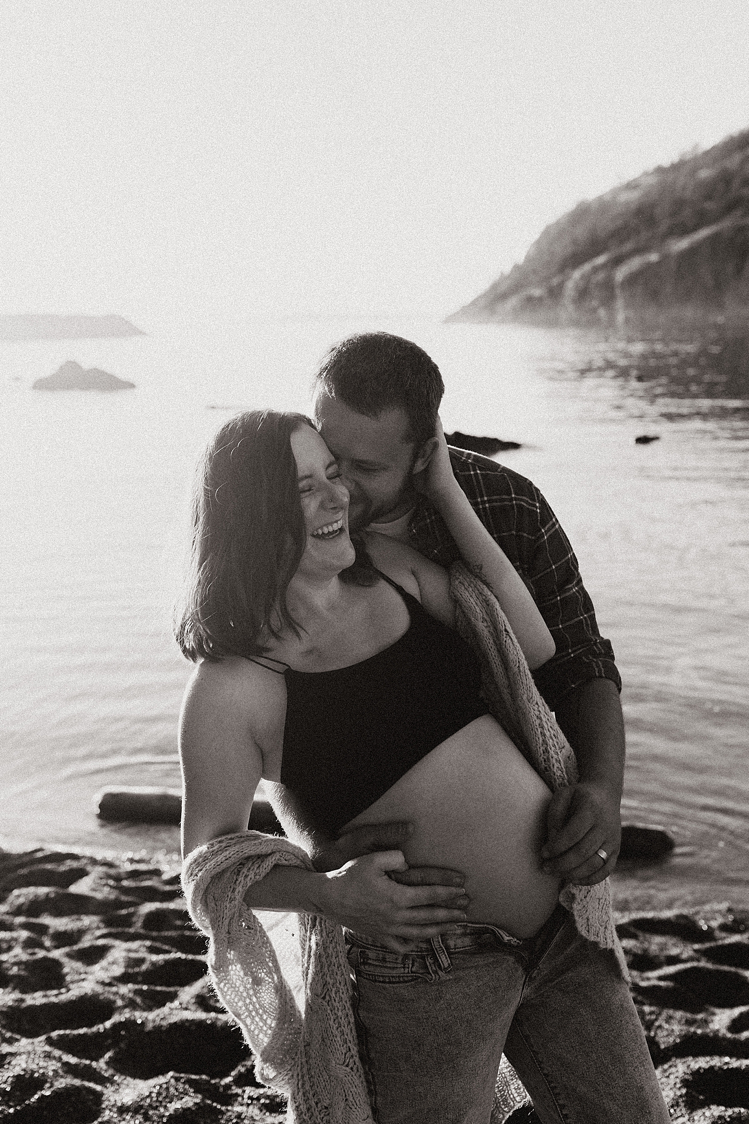 man kisses pregnant wife as she laughs by water at Rosario Beach in Washington