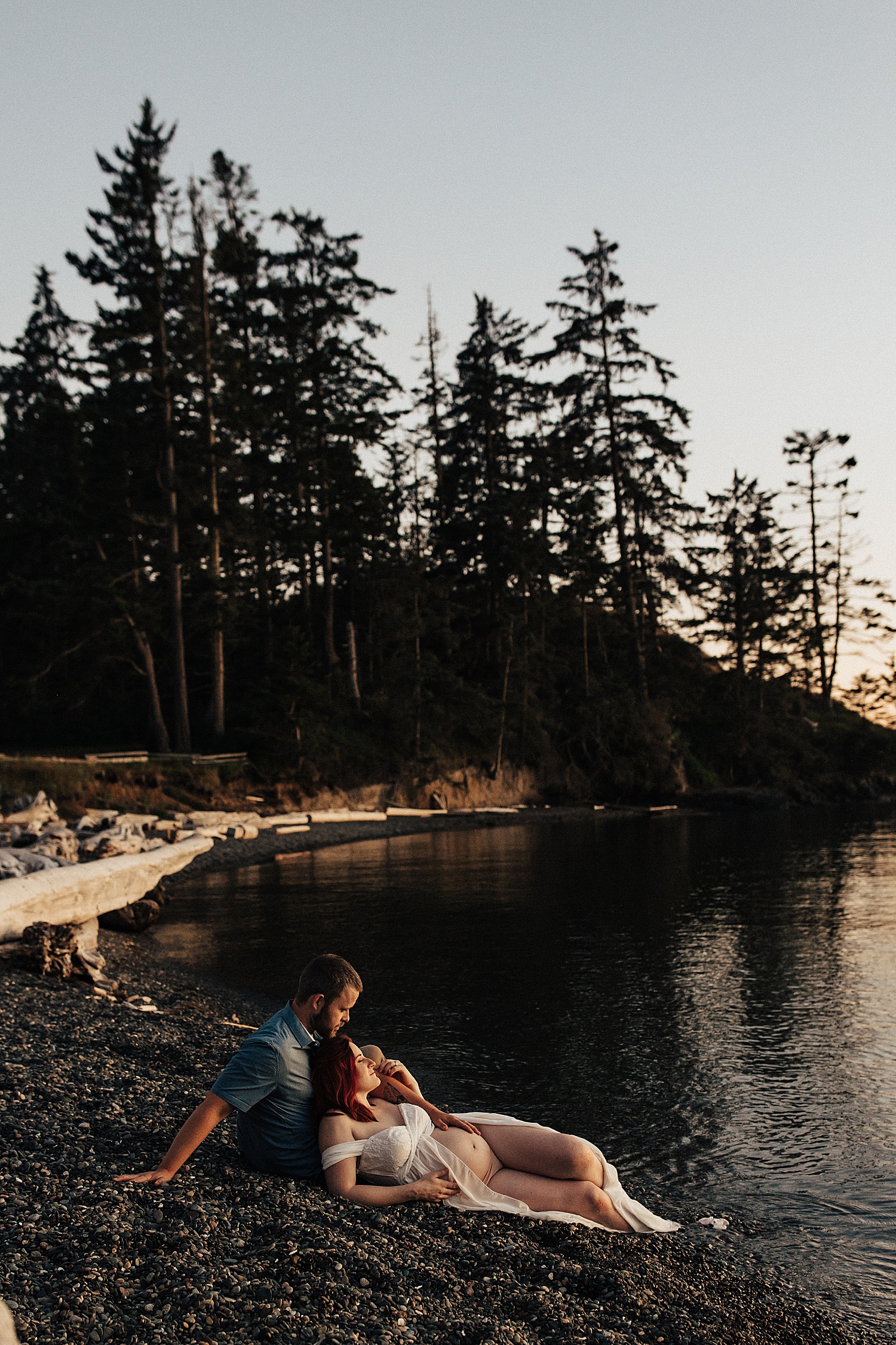 expecting mom lies on husband's lap on shoreline by destination photographer